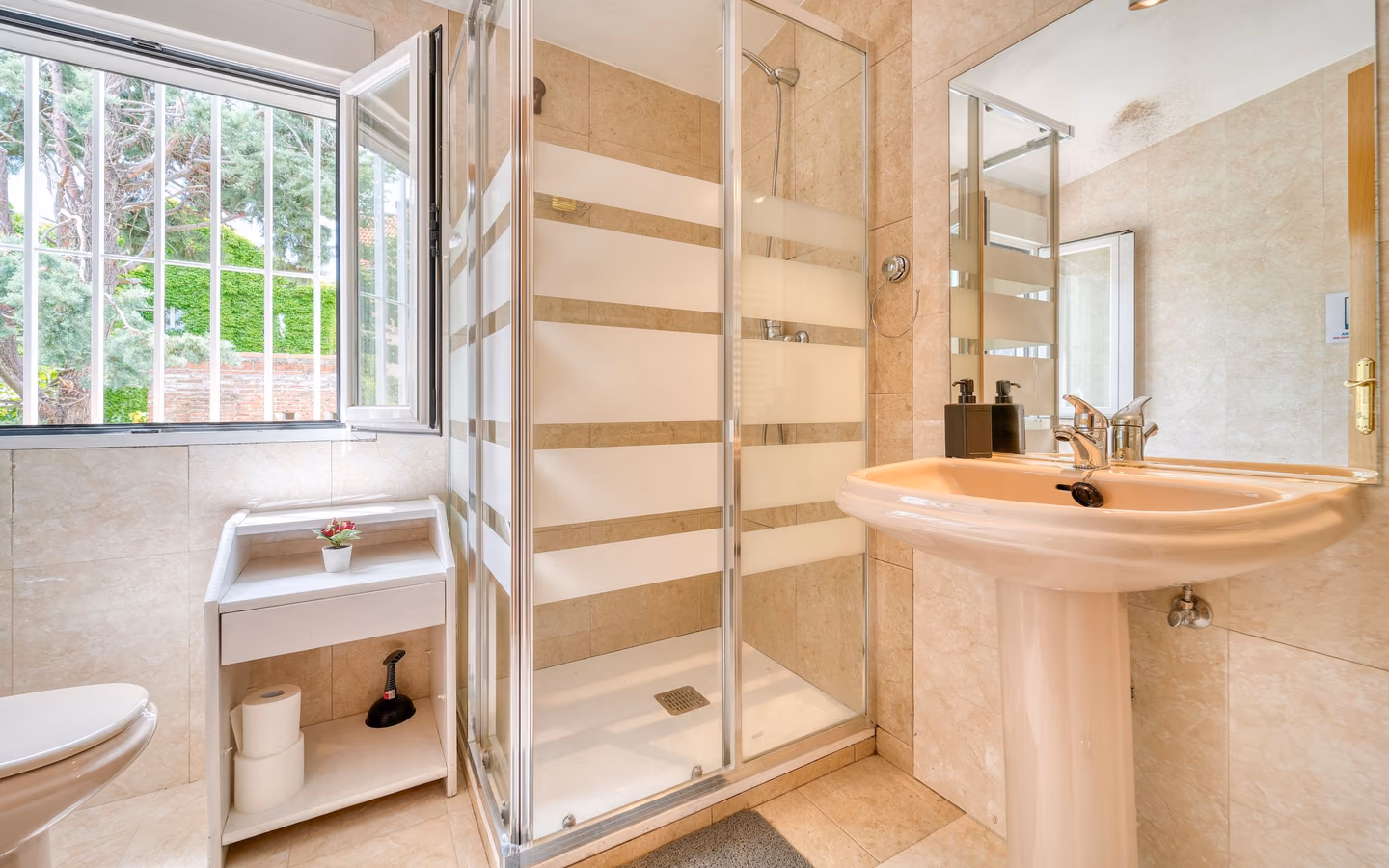 Naturally lit modern bathroom with a glass shower, pale tile, and greenery