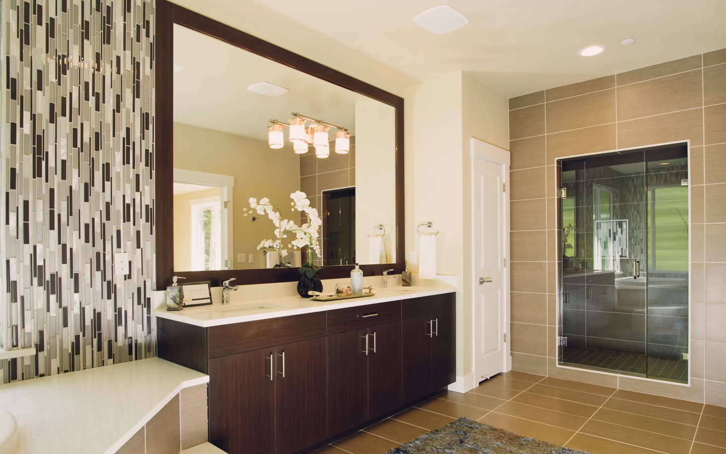 Modern bathroom with a dark wood vanity, glass shower, large mirror, and warm tile