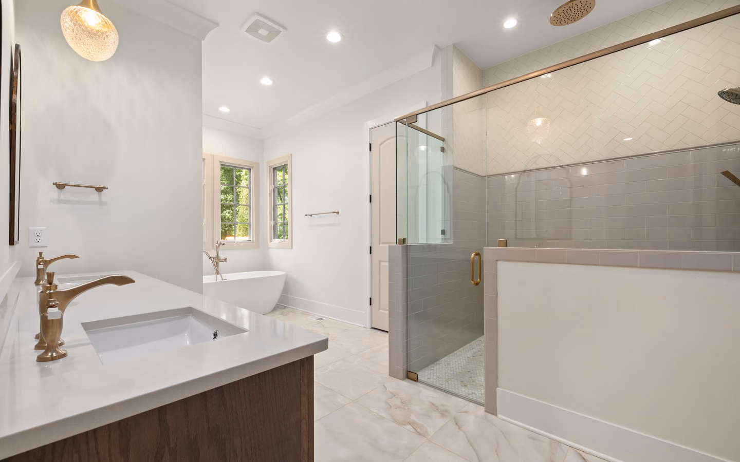Bright modern bathroom with a glass shower, bathtub, warm wood vanity, and gold accents